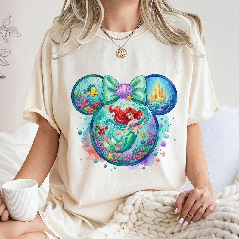 May include: Cream-coloured t-shirt with a colourful graphic of Ariel from The Little Mermaid. The design is in the shape of Mickey Mouse ears, with Ariel in the centre, surrounded by sea creatures and a castle. A large teal bow and seashell complete the design.