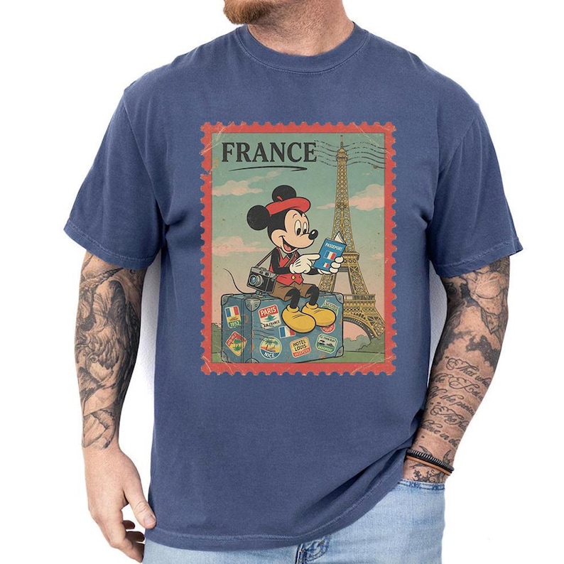 Op de afbeelding: Marineblauw T-shirt met een vintage afbeelding van Mickey Mouse voor de Eiffeltoren. De afbeelding bevat het woord "FRANCE" en een koffer met reisstickers. Mickey draagt een rode hoed en gele schoenen.