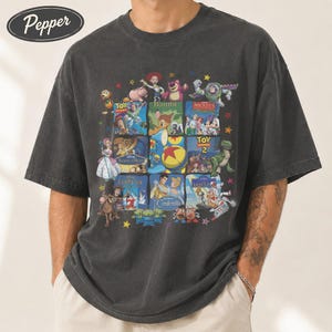 May include: Dark gray t-shirt featuring a collage of Disney movie posters, including Toy Story, Bambi, and Cinderella. The design is printed on a short-sleeved, crew neck shirt. The shirt has a vintage, faded look.