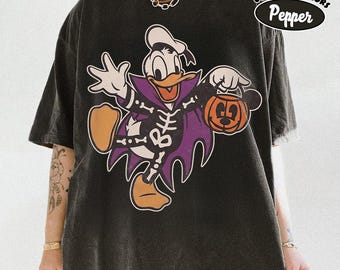 May include: A charcoal black short-sleeve t-shirt featuring a graphic of Donald Duck dressed in a skeleton costume with a purple cape, holding an orange jack-o'-lantern bucket. The text Comfort Colors Pepper is printed in the upper right corner.
