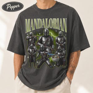 Puede incluir: Camiseta gris oscuro con un gr&aacute;fico de m&uacute;ltiples personajes Mandalorianos y el texto verde "MANDALORIAN". El dise&ntilde;o incluye una representaci&oacute;n de Grogu. La camiseta tiene un aspecto vintage y desgastado.