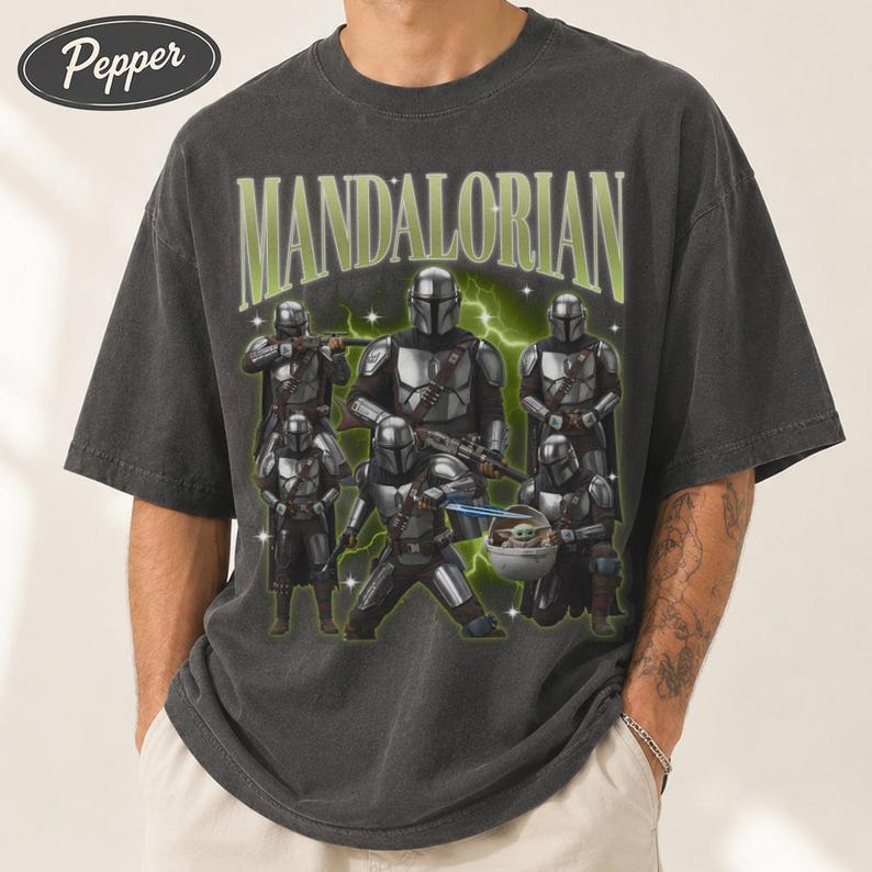 Puede incluir: Camiseta gris oscuro con un gr&aacute;fico de m&uacute;ltiples personajes Mandalorianos y el texto verde "MANDALORIAN". El dise&ntilde;o incluye una representaci&oacute;n de Grogu. La camiseta tiene un aspecto vintage y desgastado.