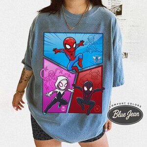 May include: Blue graphic t-shirt featuring a comic-style design with Spider-Man and other characters. The design is divided into four panels with a blue, red, and purple color scheme. The shirt has the text "Comfort Colors Blue Jean".