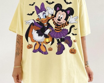 May include: A pale yellow t-shirt featuring a graphic print of Daisy Duck dressed as a vampire with a cape and lantern, and Minnie Mouse wearing a striped shirt and purple skirt. The design includes black bats and orange jack-o'-lantern pumpkins.