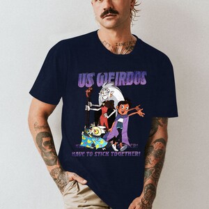 Op de afbeelding: Een donkerblauw t-shirt met een grafische print van personages uit de animatieserie "The Owl House". De grafiek toont de personages Eda, Luz en King. De tekst "US WEIRDOS" is boven de personages gedrukt, en de tekst "HAVE TO STICK TOGETHER!" is onder de personages gedrukt.