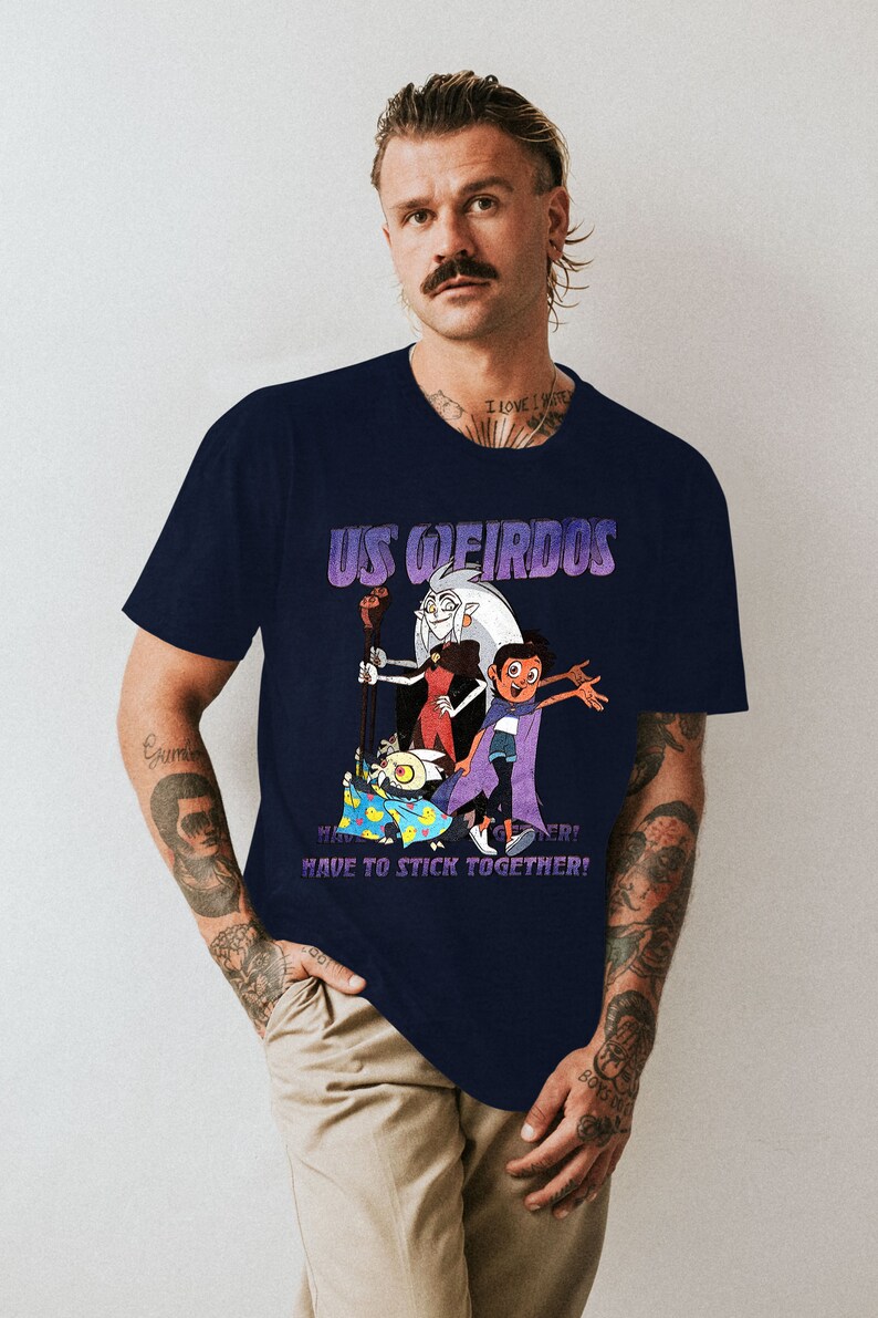 Op de afbeelding: Een donkerblauw t-shirt met een grafische print van personages uit de animatieserie "The Owl House". De grafiek toont de personages Eda, Luz en King. De tekst "US WEIRDOS" is boven de personages gedrukt, en de tekst "HAVE TO STICK TOGETHER!" is onder de personages gedrukt.