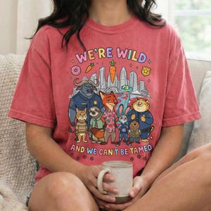 K&ouml;nnte beinhalten: Ein rot-orangefarbenes T-Shirt mit einer Cartoon-Grafik von Tierfiguren und einer Skyline. Der Text "WE'RE WILD AND WE CAN'T BE TAMED" ist oberhalb und unterhalb der Figuren aufgedruckt. Das Shirt ist aus einem weichen Material.