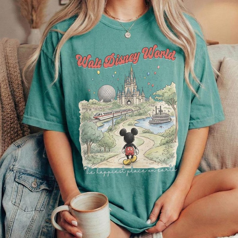 May include: Teal-colored t-shirt featuring a colorful illustration of Walt Disney World. The graphic includes the Magic Kingdom castle, a monorail, and Mickey Mouse. The text "Walt Disney World" and "The happiest place on earth" are also visible.
