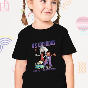Op de afbeelding: Een zwart T-shirt met een grafische print van personages uit de animatieserie "The Owl House". De grafiek toont de personages Luz, Eda en King. De tekst "US WEIRDOS HAVE TO STICK TOGETHER!" is op het shirt gedrukt.