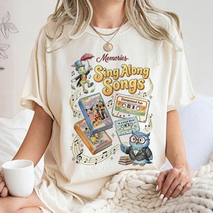 May include: Cream-colored t-shirt featuring the text "Memories Sing Along Songs" with illustrations of a cartoon owl, a cricket, cassette tapes, and VHS tapes. The design includes musical notes and a retro aesthetic.