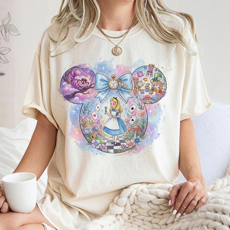 May include: Cream-colored t-shirt featuring a colorful illustration of Alice in Wonderland. The design includes the Cheshire Cat, the Mad Hatter's tea party, and Alice holding a key. A large blue bow with a clock is at the center of the design.