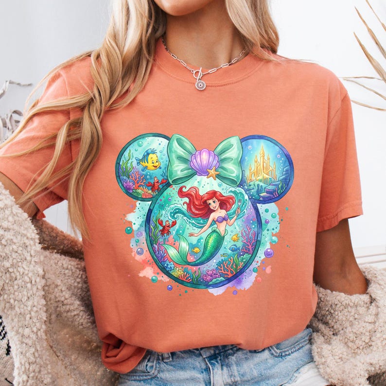 May include: Coral-coloured t-shirt featuring a colourful graphic of a mermaid inside a Mickey Mouse-shaped design. The design includes a teal bow, a seashell, and underwater scenes with fish and a castle. The shirt has short sleeves.