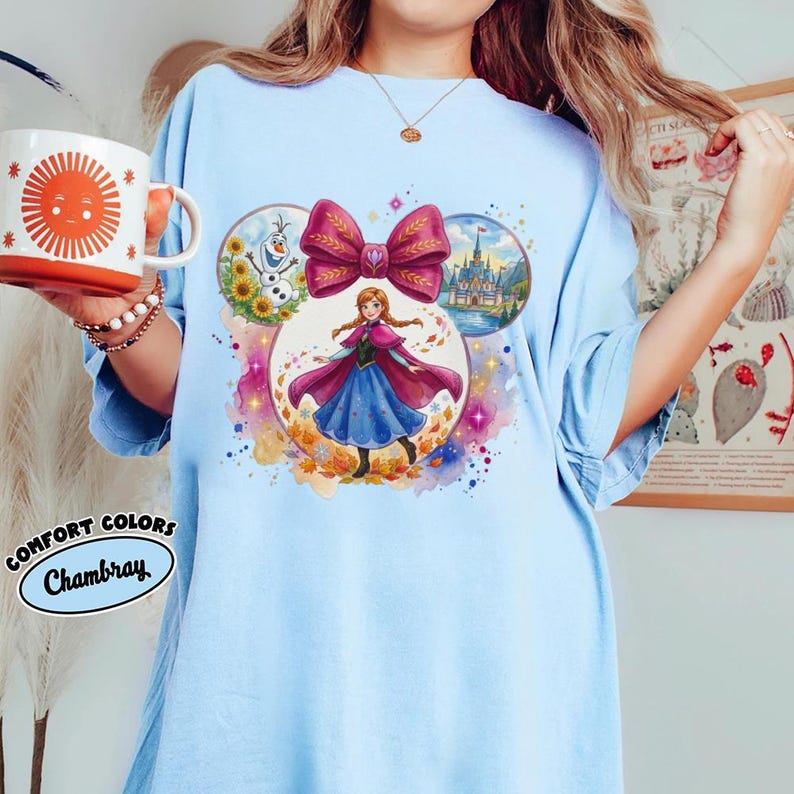 Puede incluir: Camiseta azul claro con un gr&aacute;fico colorido de una princesa, un gran lazo rosa, un castillo, girasoles y un mu&ntilde;eco de nieve. La camiseta tambi&eacute;n tiene las palabras "Comfort Colors Chambray" impresas.