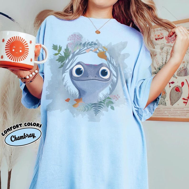 Puede incluir: Camiseta azul claro Comfort Colors Chambray con un dise&ntilde;o de salamandra de dibujos animados. La salamandra tiene ojos grandes y redondos y est&aacute; rodeada por una corona de hojas y flores. Una taza naranja a juego con un dise&ntilde;o de sol se sostiene en la mano izquierda.
