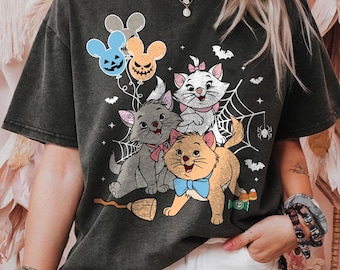 May include: Dark gray t-shirt featuring a Halloween-themed graphic of three cartoon cats, bats, spiderwebs, and balloons. The cats are in various colors, with pink and blue bows. A broom and candy corn are also included in the design.