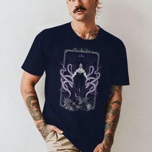 May include: A navy blue t-shirt with a graphic of Ursula, the sea witch from Disney's The Little Mermaid. The graphic is in a vintage tarot card style with the text "The Sea Witch".