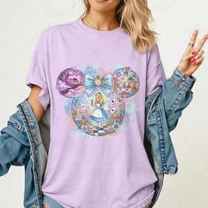 May include: A light purple t-shirt featuring a whimsical Alice in Wonderland design. The graphic includes Alice, the Cheshire Cat, and other iconic elements within a Mickey Mouse ear shape. The design is colorful and detailed, with a bow and clock.