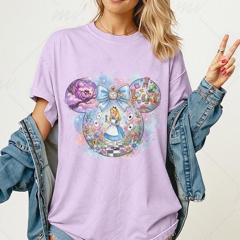 May include: A light purple t-shirt featuring a whimsical Alice in Wonderland design. The graphic includes Alice, the Cheshire Cat, and other iconic elements within a Mickey Mouse ear shape. The design is colorful and detailed, with a bow and clock.