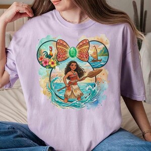 以下が含まれることがあります： 明るい紫色のTシャツ。キャラクターがパドルを持ち、海の波と熱帯の要素に囲まれたカラフルなグラフィックが特徴です。デザインはミッキーマウスの耳の形の中に配置され、上部に模様入りのリボンが付いています。