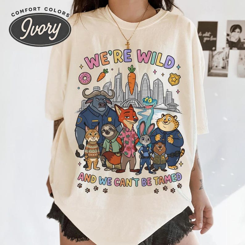 K&ouml;nnte beinhalten: Cremefarbenes T-Shirt mit einer farbenfrohen Grafik mit Zeichentieren und dem Text "WE'RE WILD AND WE CAN'T BE TAMED." Das Design umfasst eine Stadtlandschaft, Donuts, Karotten und Pfotenabdr&uuml;cke.