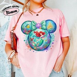 May include: Pink t-shirt with a colourful graphic of Ariel the mermaid within a Mickey Mouse head shape. The design includes a teal bow, a golden castle, and other sea creatures. The text "Comfort Colors Blossom" is in a white oval on the top left.