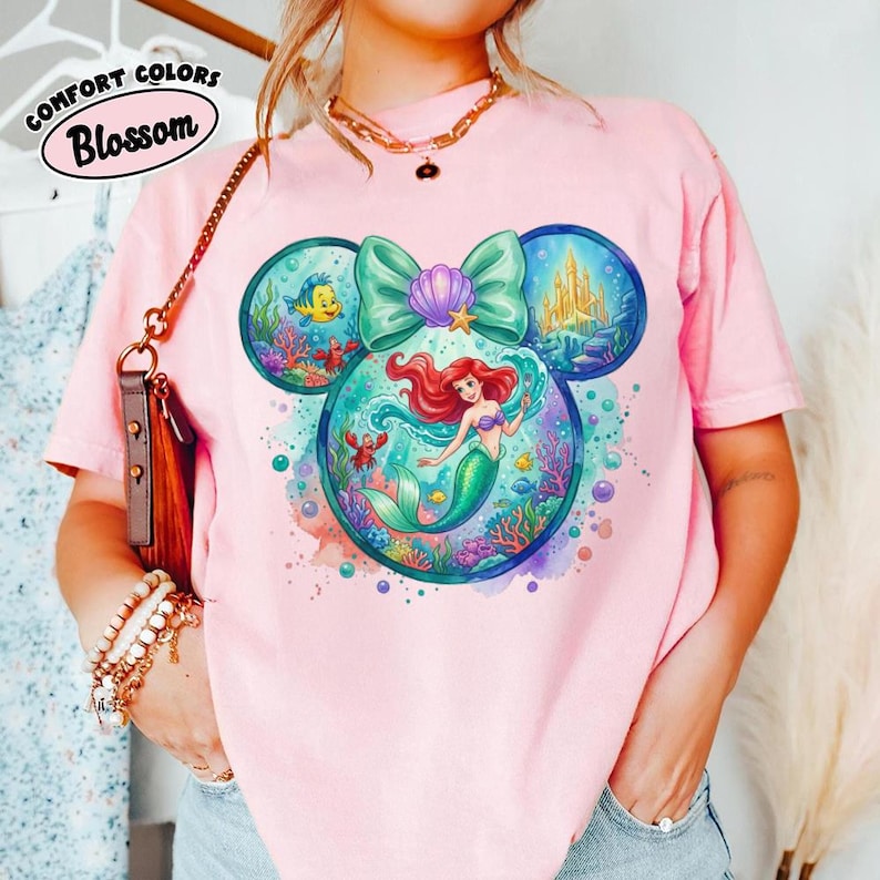 May include: Pink t-shirt with a colourful graphic of Ariel the mermaid within a Mickey Mouse head shape. The design includes a teal bow, a golden castle, and other sea creatures. The text "Comfort Colors Blossom" is in a white oval on the top left.