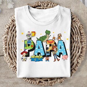 Puede incluir: Camiseta blanca con un gr&aacute;fico colorido de los personajes de Toy Story que deletrean "PAPA".