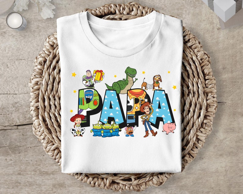 Puede incluir: Camiseta blanca con un gr&aacute;fico colorido de los personajes de Toy Story que deletrean "PAPA".