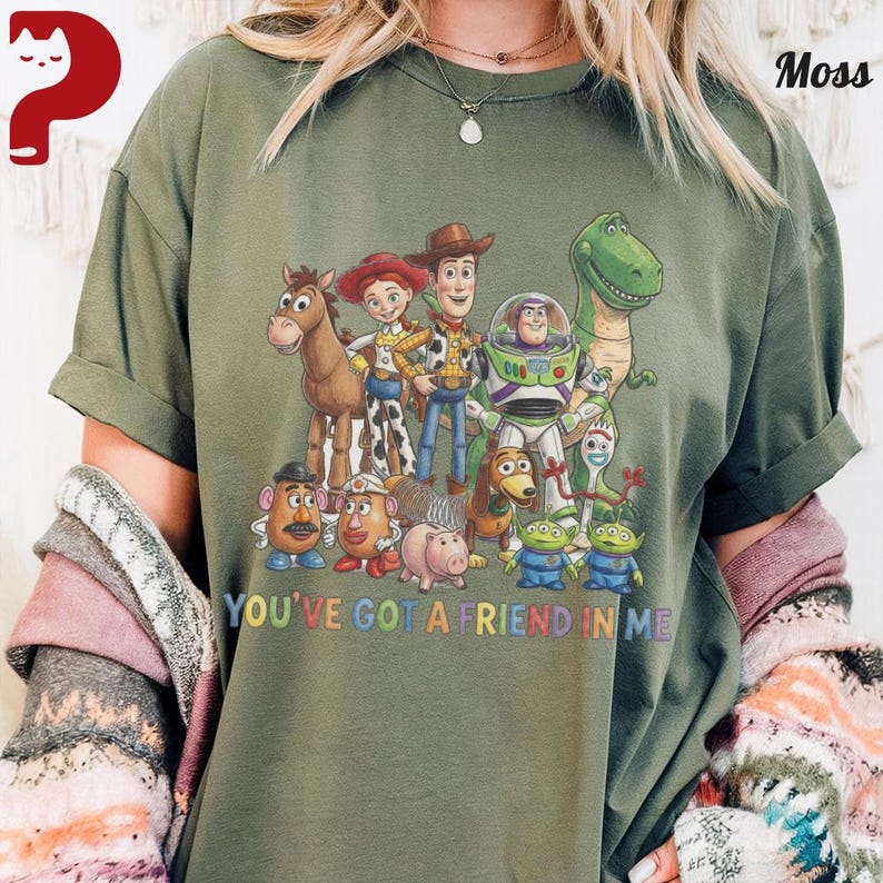 May include: Olive green t-shirt featuring a colorful graphic of characters from the animated film "Toy Story." The design includes Woody, Buzz Lightyear, and other characters, with the text "YOU'VE GOT A FRIEND IN ME" in a rainbow font.
