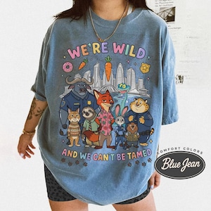 K&ouml;nnte beinhalten: Blaues Comfort Colors T-Shirt mit einer Grafik von Zeichentrickfiguren und dem Text "WE'RE WILD AND WE CAN'T BE TAMED". Das Design umfasst eine Stadtlandschaft, Donuts und Karotten.