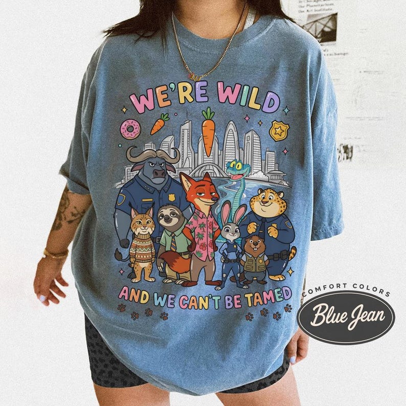 K&ouml;nnte beinhalten: Blaues Comfort Colors T-Shirt mit einer Grafik von Zeichentrickfiguren und dem Text "WE'RE WILD AND WE CAN'T BE TAMED". Das Design umfasst eine Stadtlandschaft, Donuts und Karotten.