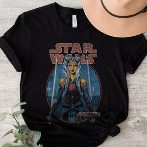 Op de afbeelding: Zwart T-shirt met een vintage-stijl afbeelding van Ahsoka Tano uit Star Wars. De afbeelding toont Ahsoka met haar Togruta-make-up en blauwe en witte hoofdtooi, die twee lichtzwaarden vasthoudt. De tekst "STAR WARS" staat boven de afbeelding, en "AHSOKA" eronder.