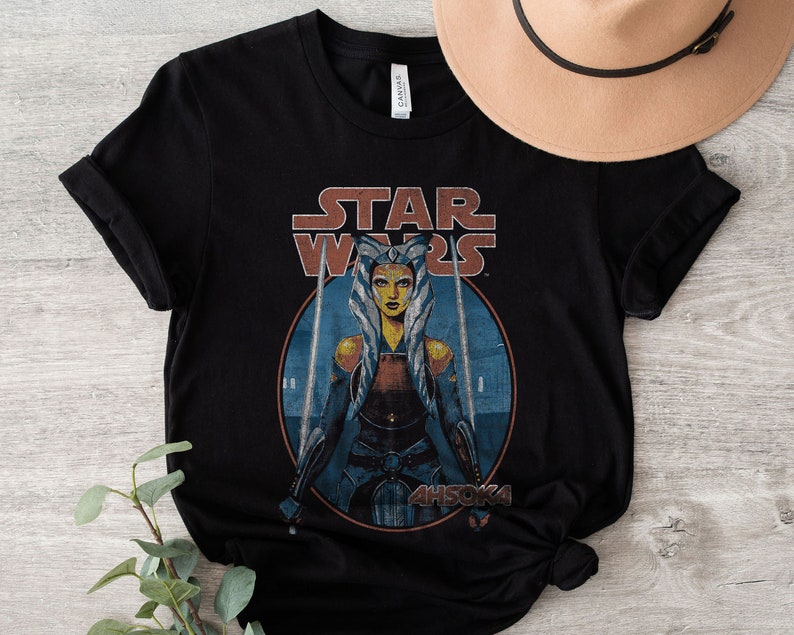 Op de afbeelding: Zwart T-shirt met een vintage-stijl afbeelding van Ahsoka Tano uit Star Wars. De afbeelding toont Ahsoka met haar Togruta-make-up en blauwe en witte hoofdtooi, die twee lichtzwaarden vasthoudt. De tekst "STAR WARS" staat boven de afbeelding, en "AHSOKA" eronder.