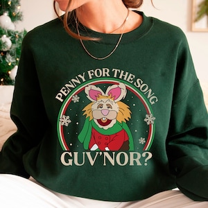 May include: A green sweatshirt with a white graphic of a cartoon rabbit wearing a red and green outfit. The graphic says "Penny for the song Guv'nor?" with snowflakes around the rabbit.