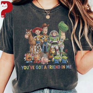 May include: Dark gray t-shirt featuring a colorful graphic of characters from the animated film "Toy Story." The design includes Woody, Buzz Lightyear, and other beloved characters, with the text "YOU'VE GOT A FRIEND IN ME" below.