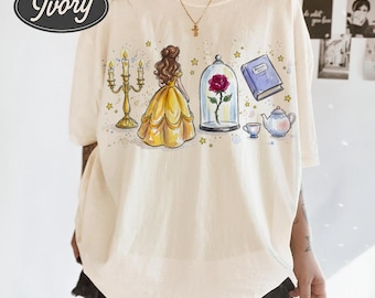 May include: Ivory-colored t-shirt with a whimsical illustration featuring a yellow gown, a rose in a glass dome, a blue book, a teapot, and a candelabra. The shirt has the words "Comfort Colors" and "Ivory" in a circular design.