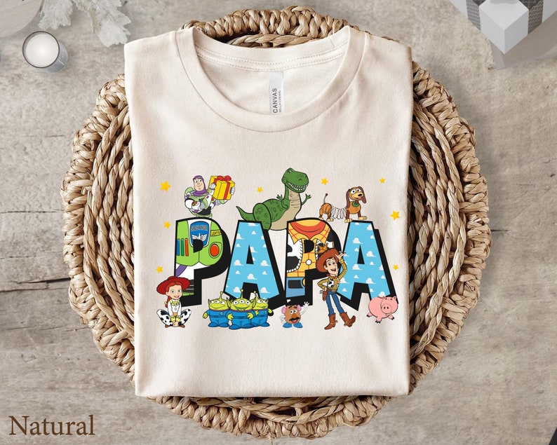 Puede incluir: Una camiseta beige con un gr&aacute;fico de los personajes de Toy Story formando la palabra "PAPA". Los personajes incluyen a Woody, Buzz Lightyear, Rex y Jessie.