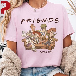 May include: A light pink t-shirt featuring the word "FRIENDS" in brown letters above a graphic of Toy Story characters sitting on a couch. The text "SINCE 1995" is below the couch.