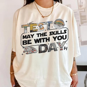 May include: Cream-colored t-shirt featuring a Star Wars-themed graphic. The design includes the text "TEST DAY" with the phrase "MAY THE SKILLS BE WITH YOU." Illustrations of Baby Yoda and the Millennium Falcon are also present.