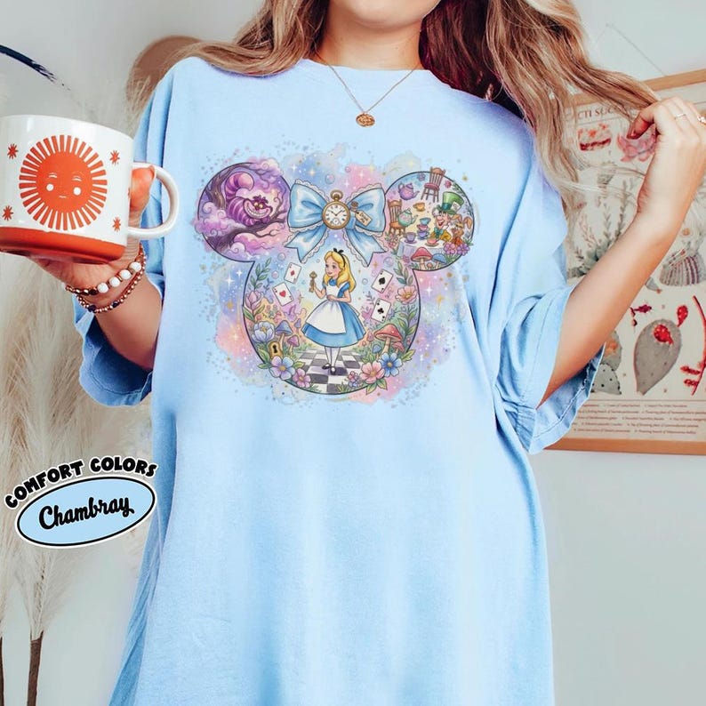 May include: Light blue t-shirt featuring a whimsical Alice in Wonderland design within a Mickey Mouse silhouette. The design includes Alice, the Cheshire Cat, and other iconic elements. A mug with a smiling sun graphic is held in the hand.