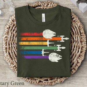 May include: A military green t-shirt featuring a rainbow stripe design and a vintage-style image of spaceships flying in formation. The text "Military Green" is visible below the image.
