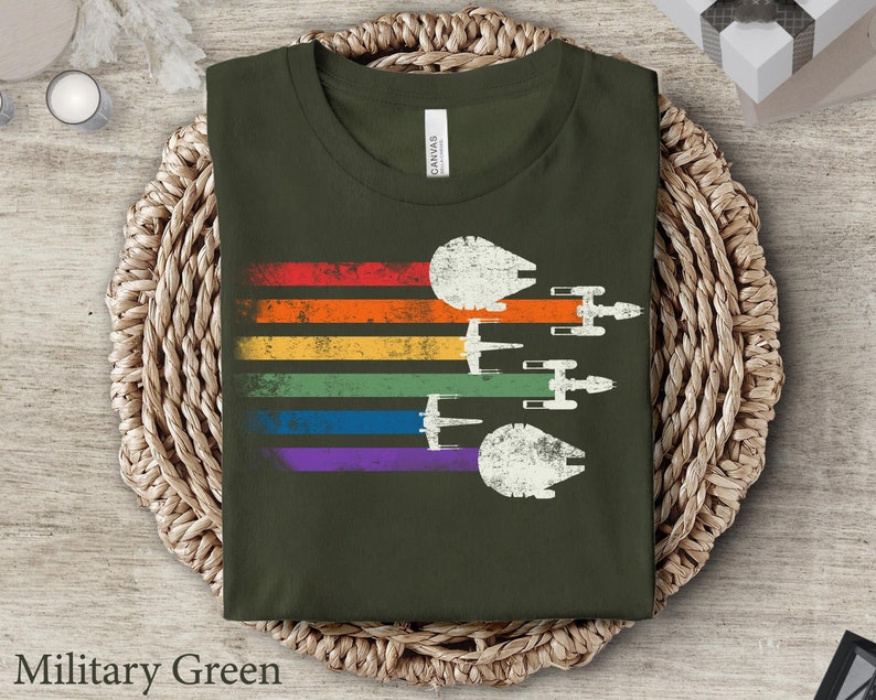 May include: A military green t-shirt featuring a rainbow stripe design and a vintage-style image of spaceships flying in formation. The text "Military Green" is visible below the image.