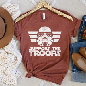 May include: A rust-colored t-shirt with a white graphic of a Star Wars Stormtrooper helmet with the text "Support the Troops" underneath.