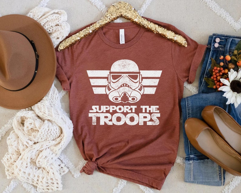 May include: A rust-colored t-shirt with a white graphic of a Star Wars Stormtrooper helmet with the text "Support the Troops" underneath.