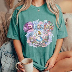 May include: Teal-colored t-shirt featuring a whimsical Alice in Wonderland design. The graphic, shaped like Mickey Mouse ears, showcases Alice, the Cheshire Cat, and other characters. A large blue bow and pocket watch are also included in the design.