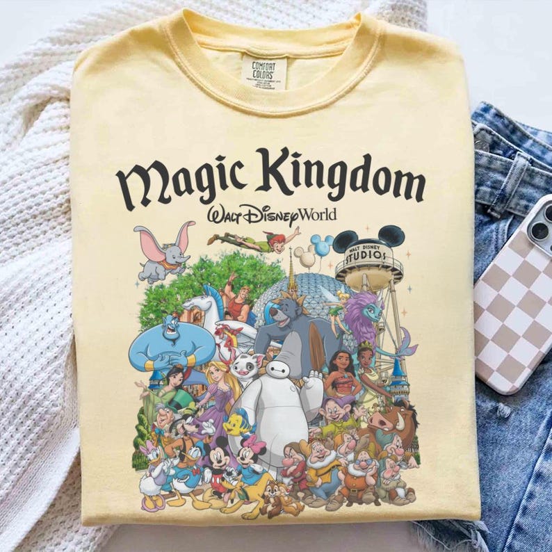 May include: A light yellow t-shirt featuring the words "Magic Kingdom" above a colorful graphic of Disney characters. The graphic includes characters like Mickey Mouse, Dumbo, and Baymax. The shirt is folded.