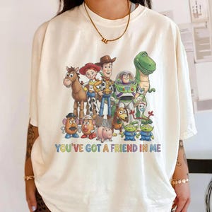 May include: Cream-colored t-shirt featuring a colorful graphic of characters from the animated film "Toy Story." The design includes Woody, Buzz Lightyear, and other characters, with the text "You've Got a Friend in Me" in a rainbow font.