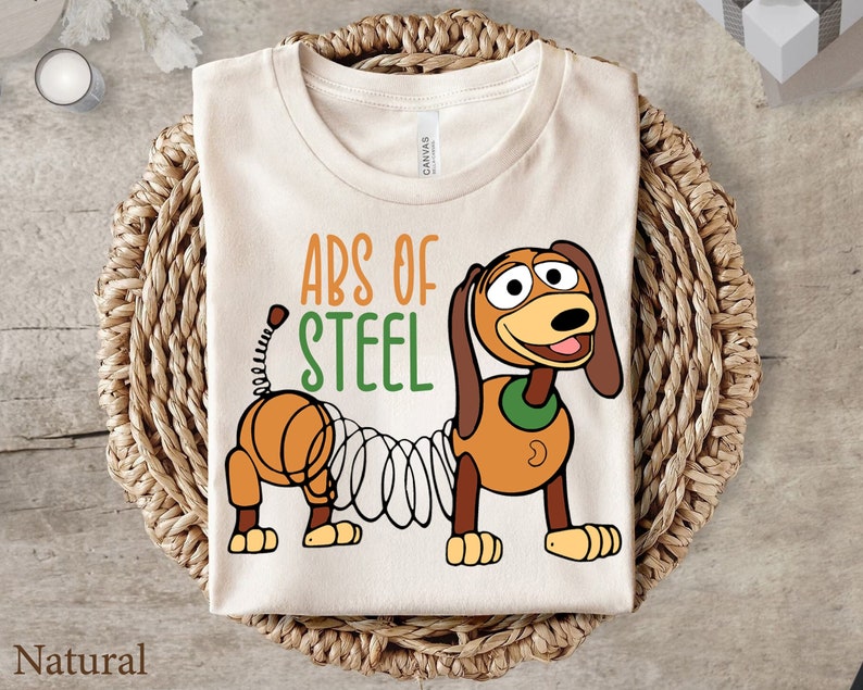 Puede incluir: Una camiseta beige con el texto "Abs of Steel" y una imagen de dibujos animados de un perro marr&oacute;n y naranja con un muelle para la cola. El perro lleva un collar verde.