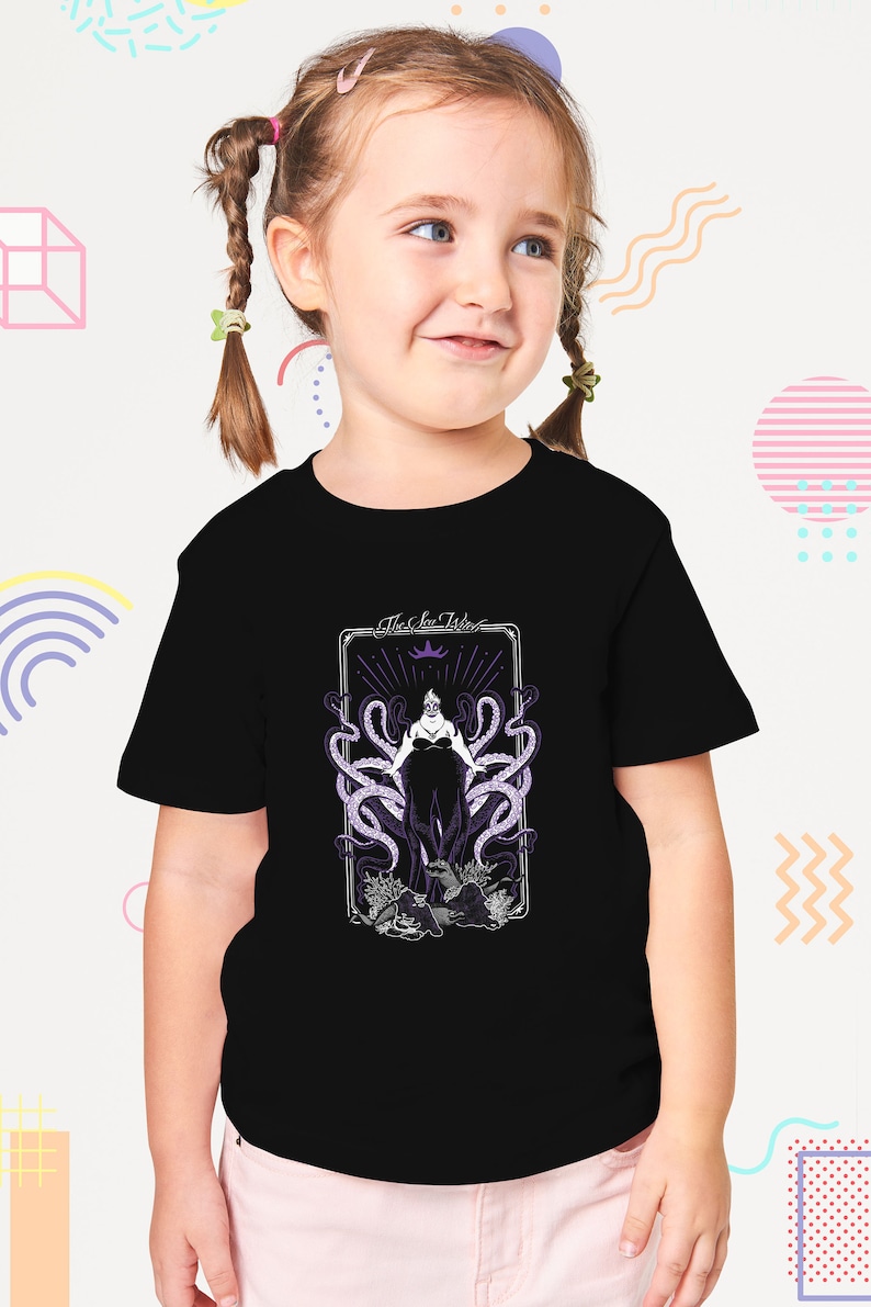 May include: A black t-shirt with a graphic of Ursula, the Sea Witch, from Disney's The Little Mermaid. The graphic is in a vintage style with a purple and white color scheme. The text "The Sea Witch" is above the graphic.