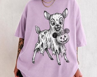 May include: A light purple short-sleeve t-shirt featuring a black and white graphic illustration of a young deer wearing a tattered ghost costume and holding a jack-o'-lantern candy bucket filled with various round treats.